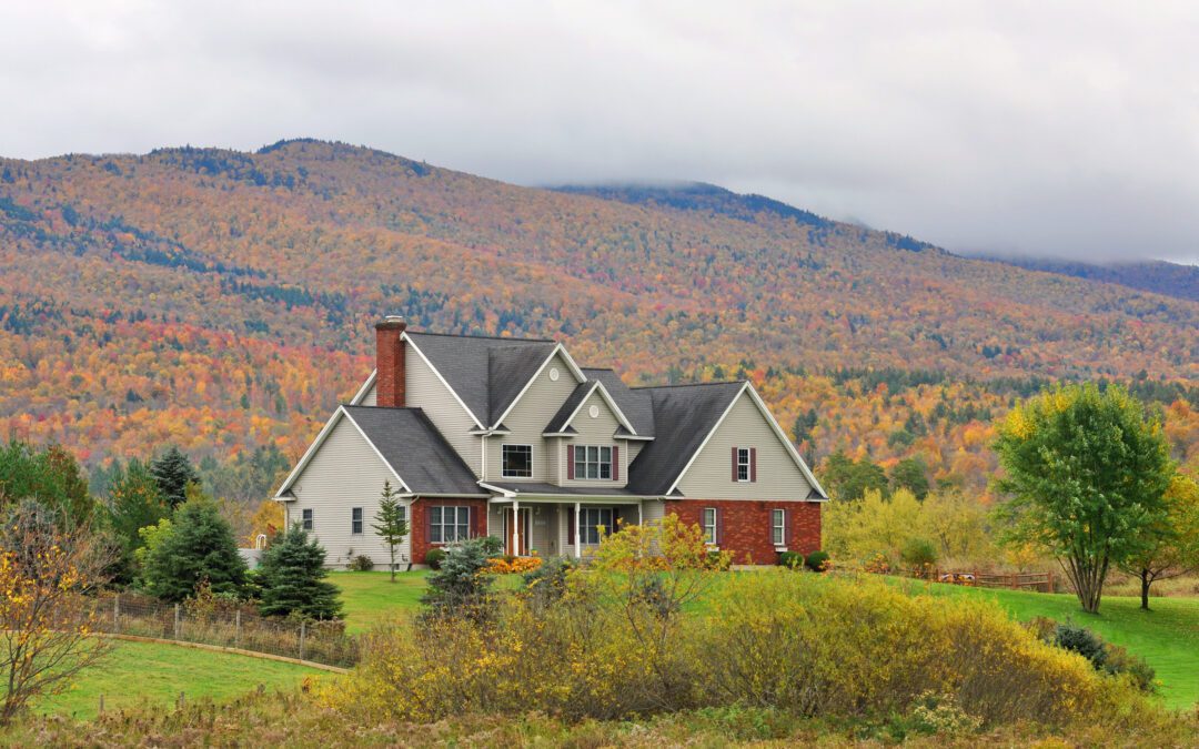 A charming house with large windows and a wraparound porch, nestled in a valley surrounded by majestic mountains and lush, vibrant trees.